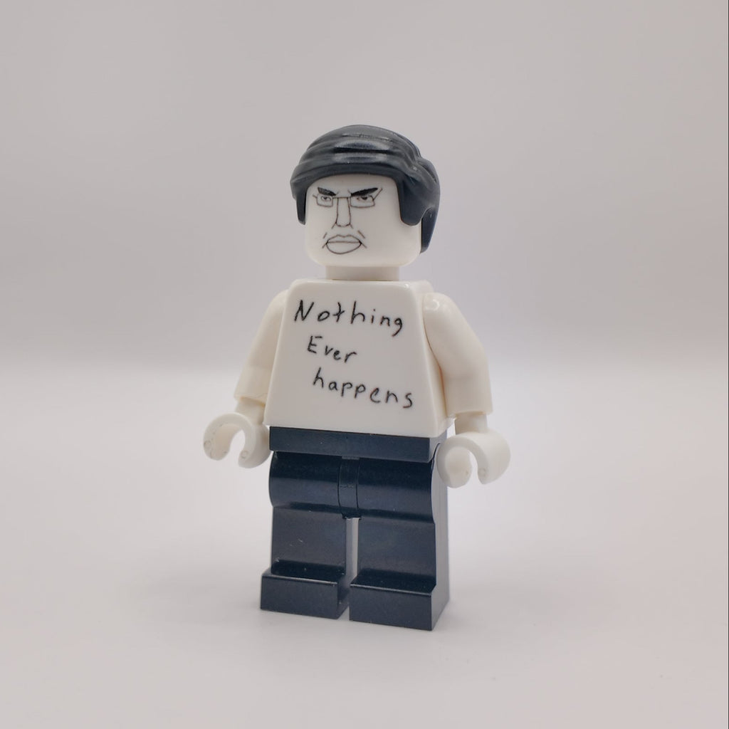 A custom LEGO minifigure with a white shirt that reads 'Nothing Ever Happens' in black handwriting, black hair, and dark blue pants, standing against a plain light gray background.