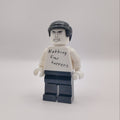 A custom LEGO minifigure with a white shirt that reads 'Nothing Ever Happens' in black handwriting, black hair, and dark blue pants, standing against a plain light gray background.