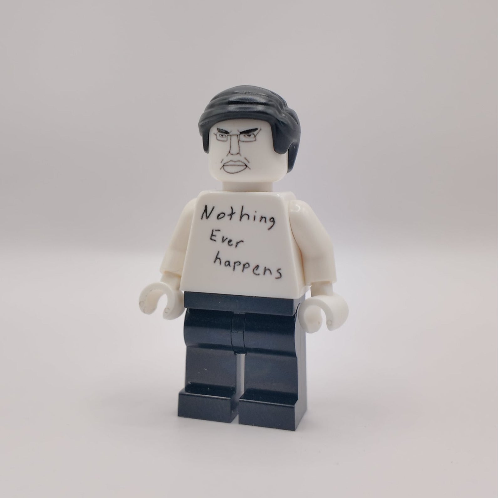 A custom LEGO minifigure with a white shirt that reads 'Nothing Ever Happens' in black handwriting, black hair, and dark blue pants, standing against a plain light gray background.