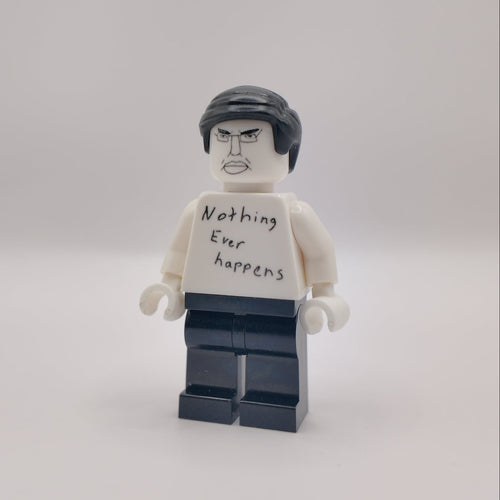A custom LEGO minifigure with a white shirt that reads 'Nothing Ever Happens' in black handwriting, black hair, and dark blue pants, standing against a plain light gray background.