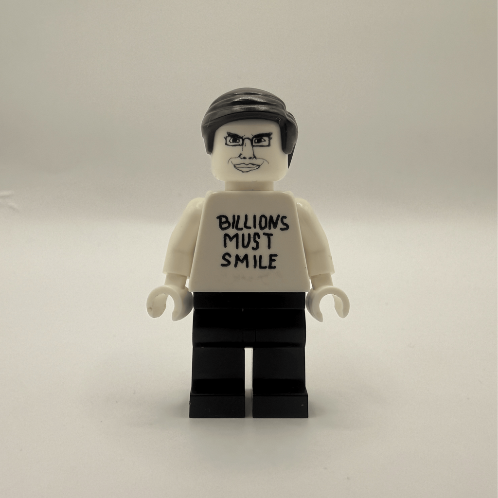 A custom Lego minifigure with black hair, a white shirt printed with the text 'BILLIONS MUST SMILE' in black, and black pants, standing against a plain light gray background.