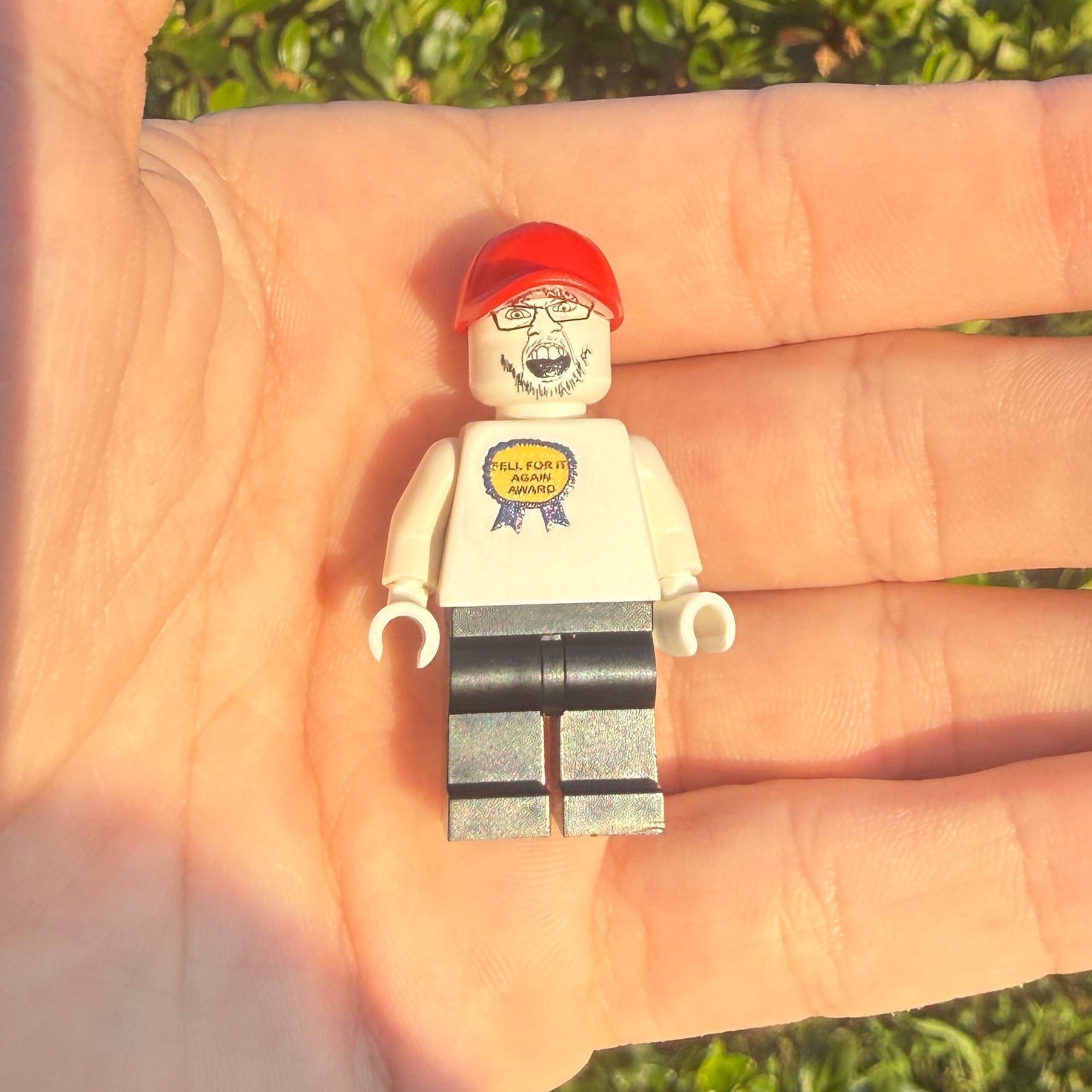 A small LEGO minifigure with a red cap, white shirt, and gray pants resting on an open hand, with green bushes in the background.