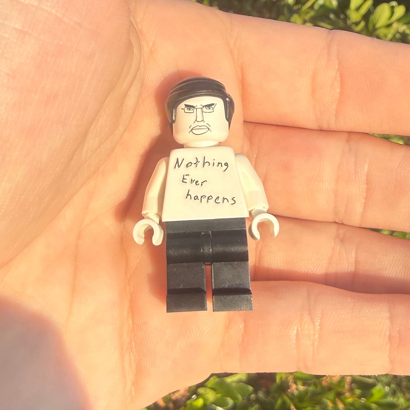 A small toy minifigure with a printed face and the text 'Nothing ever happens' on its shirt, held in the palm of a hand against a background of green outdoor foliage.