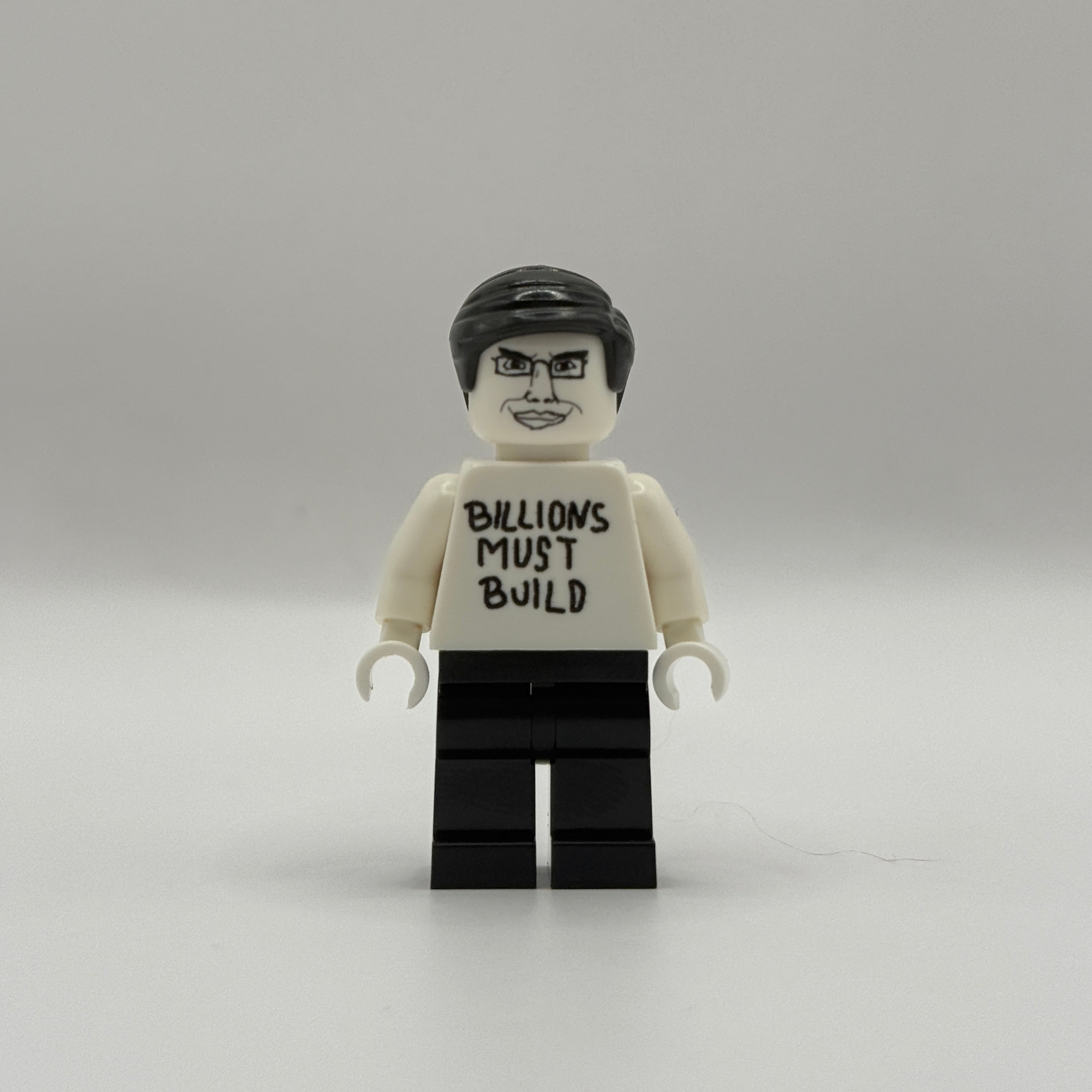 A custom-built toy minifigure resembling a LEGO figure, featuring a white torso printed with the text 'BILLIONS MUST BUILD' in black, black hair, and black legs, set against a plain light gray background.