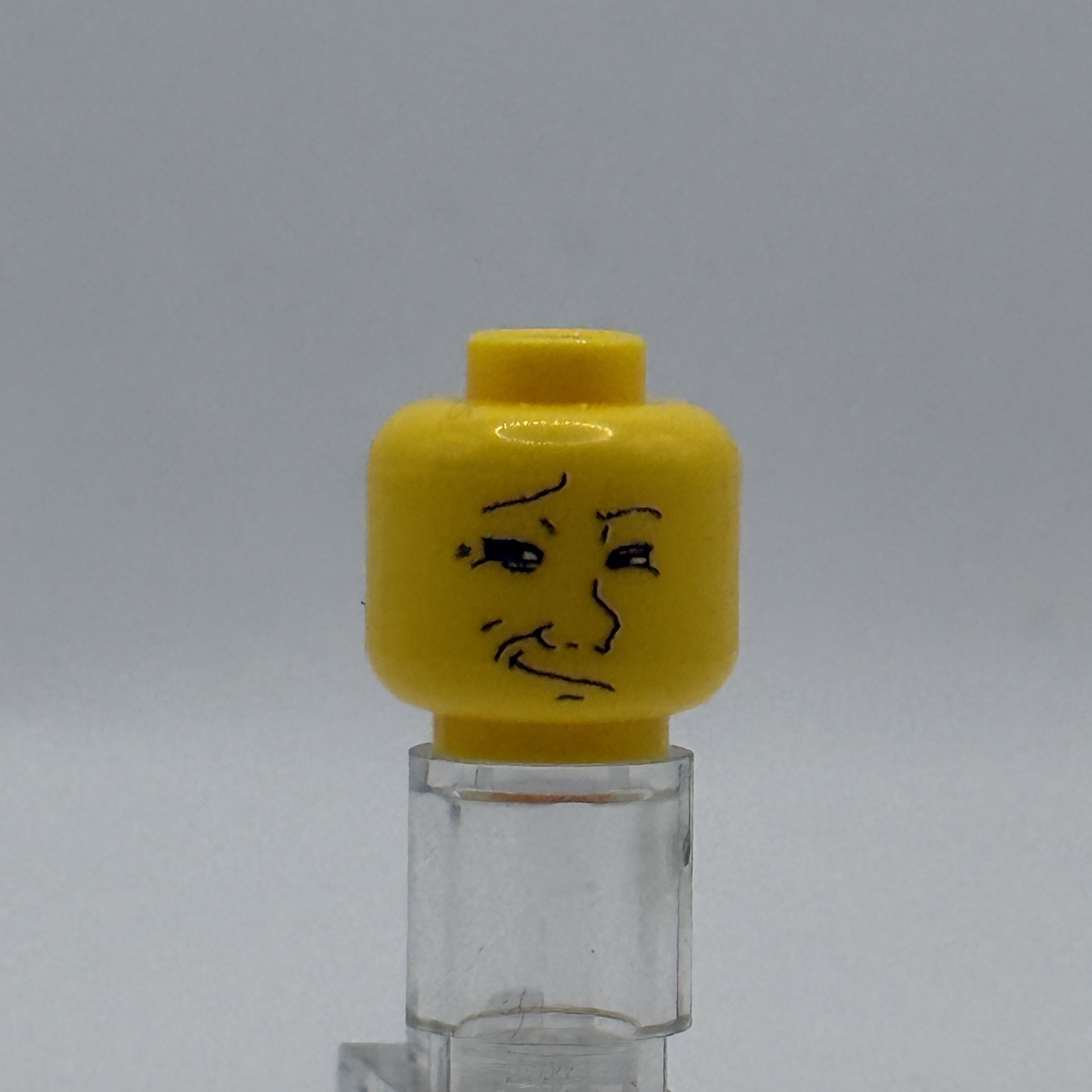 A yellow LEGO minifigure head with a drawn sad face, mounted on a clear plastic display stand against a neutral gray background.