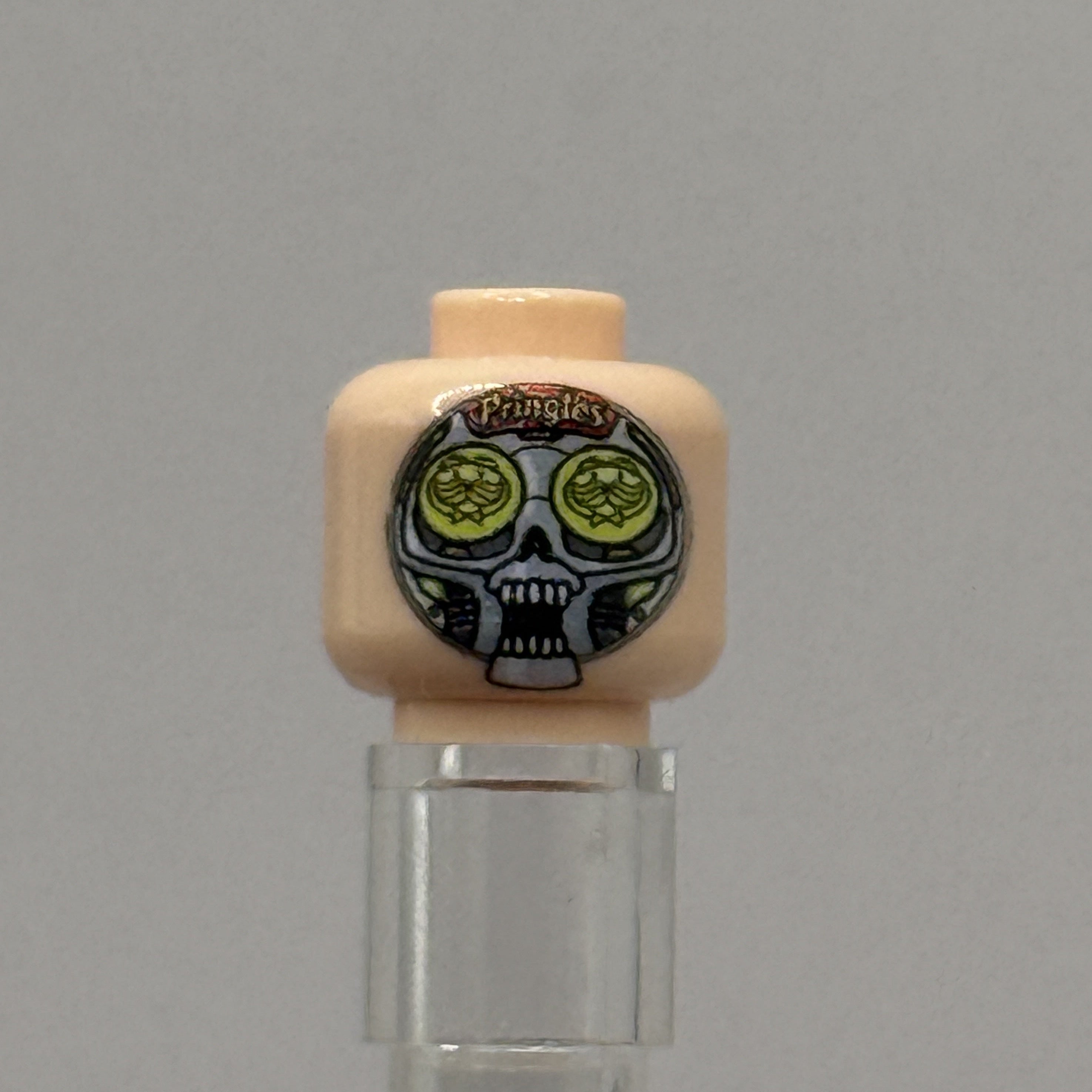 A custom-printed LEGO minifigure head with a beige skin tone, featuring a black and white skull design with green circular eyes and the text 'Pringles' on a gray background.