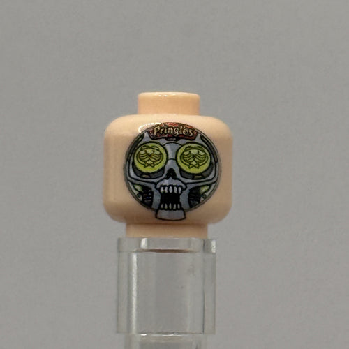 A custom-printed LEGO minifigure head with a beige skin tone, featuring a black and white skull design with green circular eyes and the text 'Pringles' on a gray background.