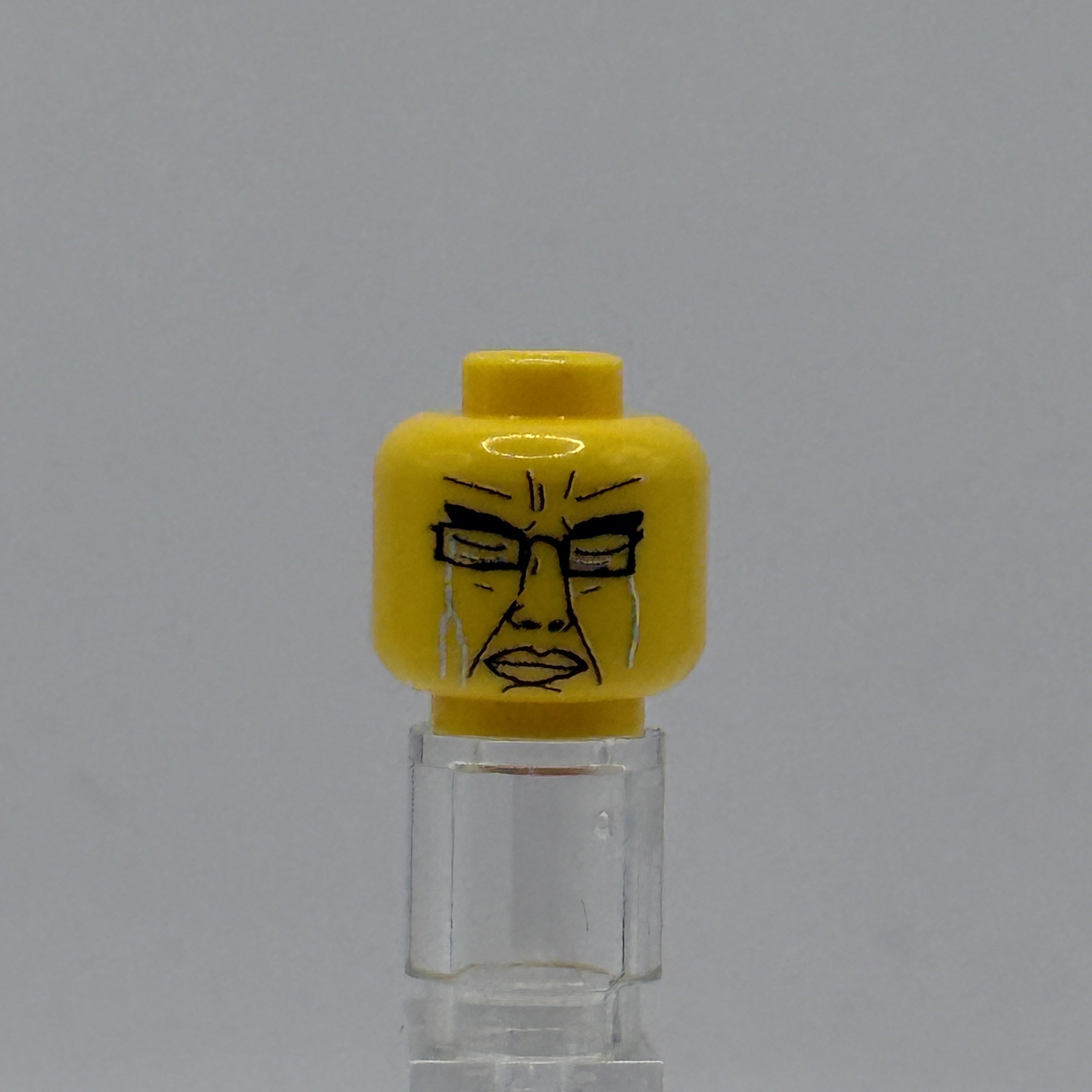 A yellow LEGO minifigure head with a black printed face, displayed on a clear acrylic stand against a neutral gray background.