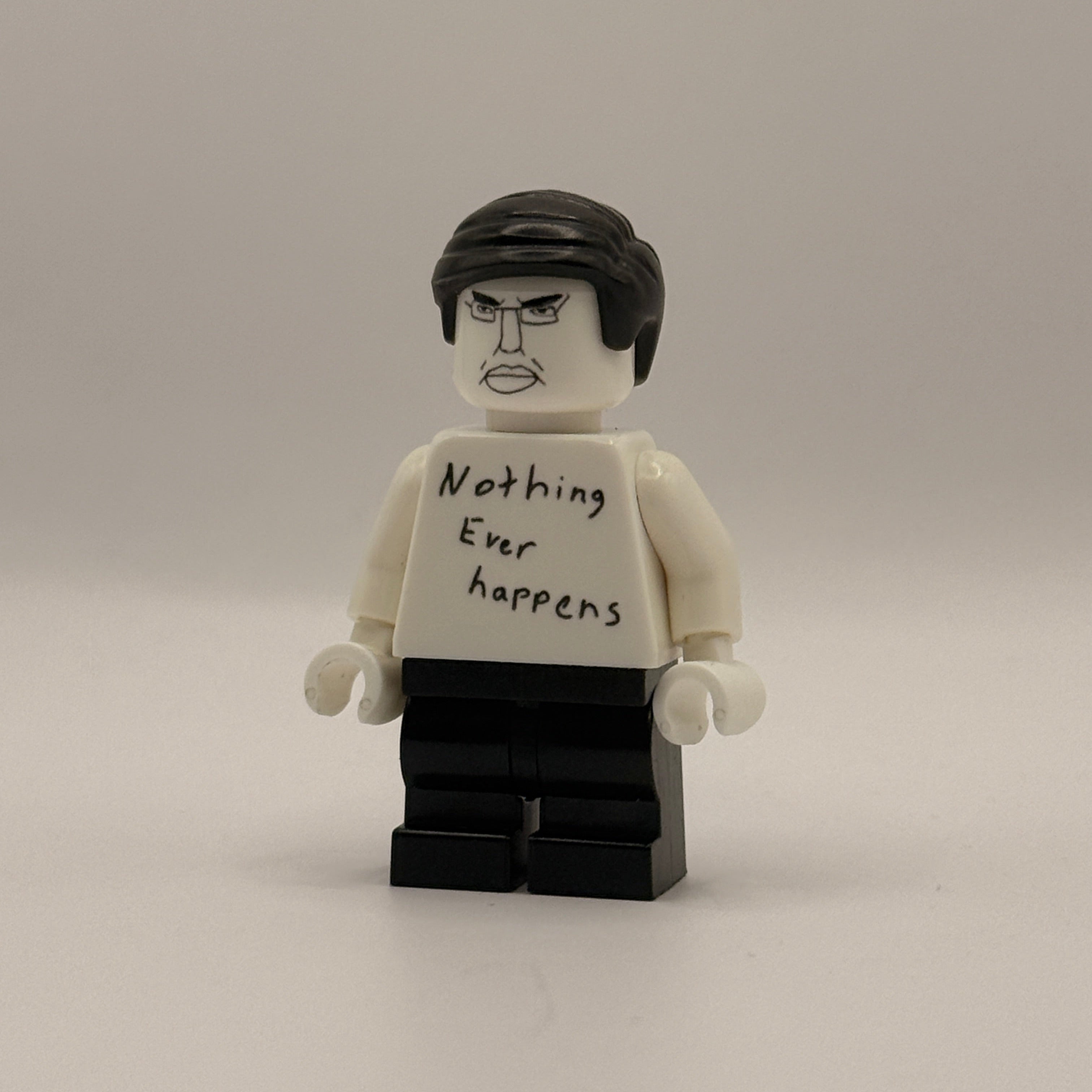 A custom LEGO minifigure with a white torso and head, black hair, and a black brick base. The torso has handwritten text reading 'Nothing ever happens' on the back.