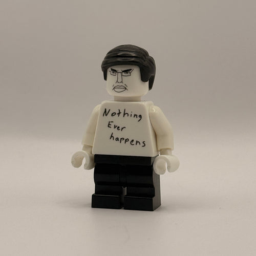 A custom LEGO minifigure with a white torso and head, black hair, and a black brick base. The torso has handwritten text reading 'Nothing ever happens' on the back.