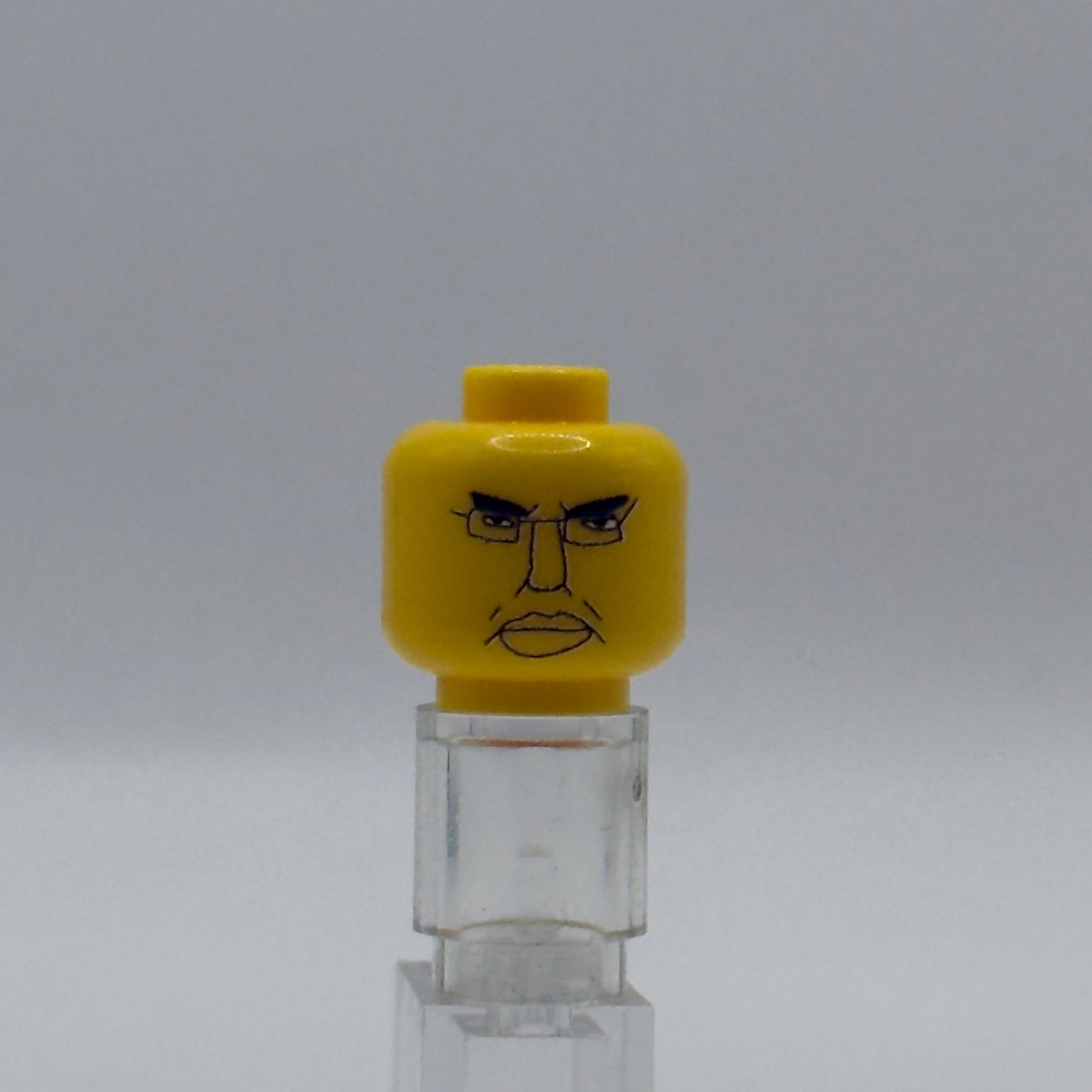 A yellow LEGO minifigure head with an angry printed face, displayed on a clear acrylic stand against a neutral gray background.