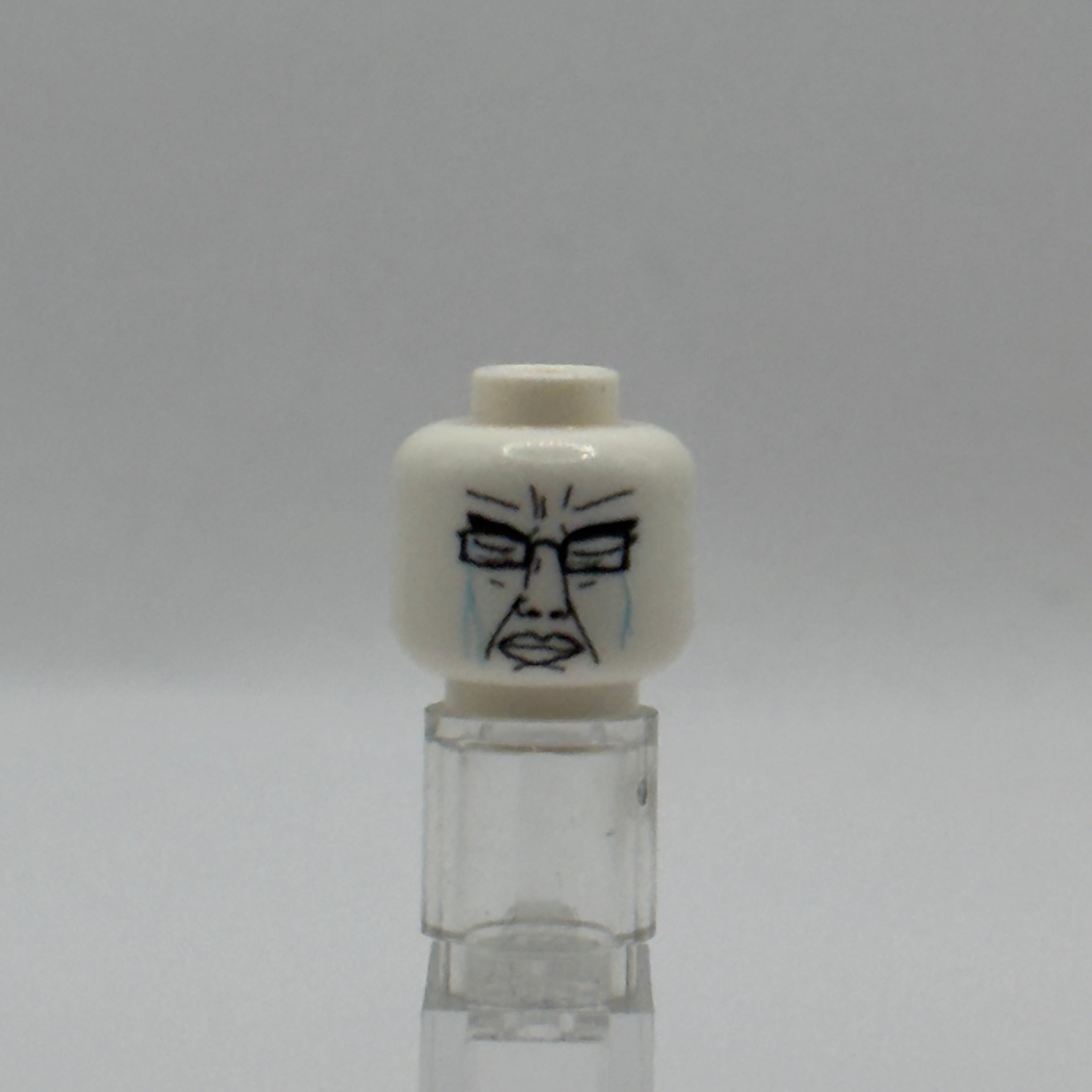A white LEGO minifigure head with a printed black and blue crying face, displayed on a clear plastic stand against a neutral gray background.