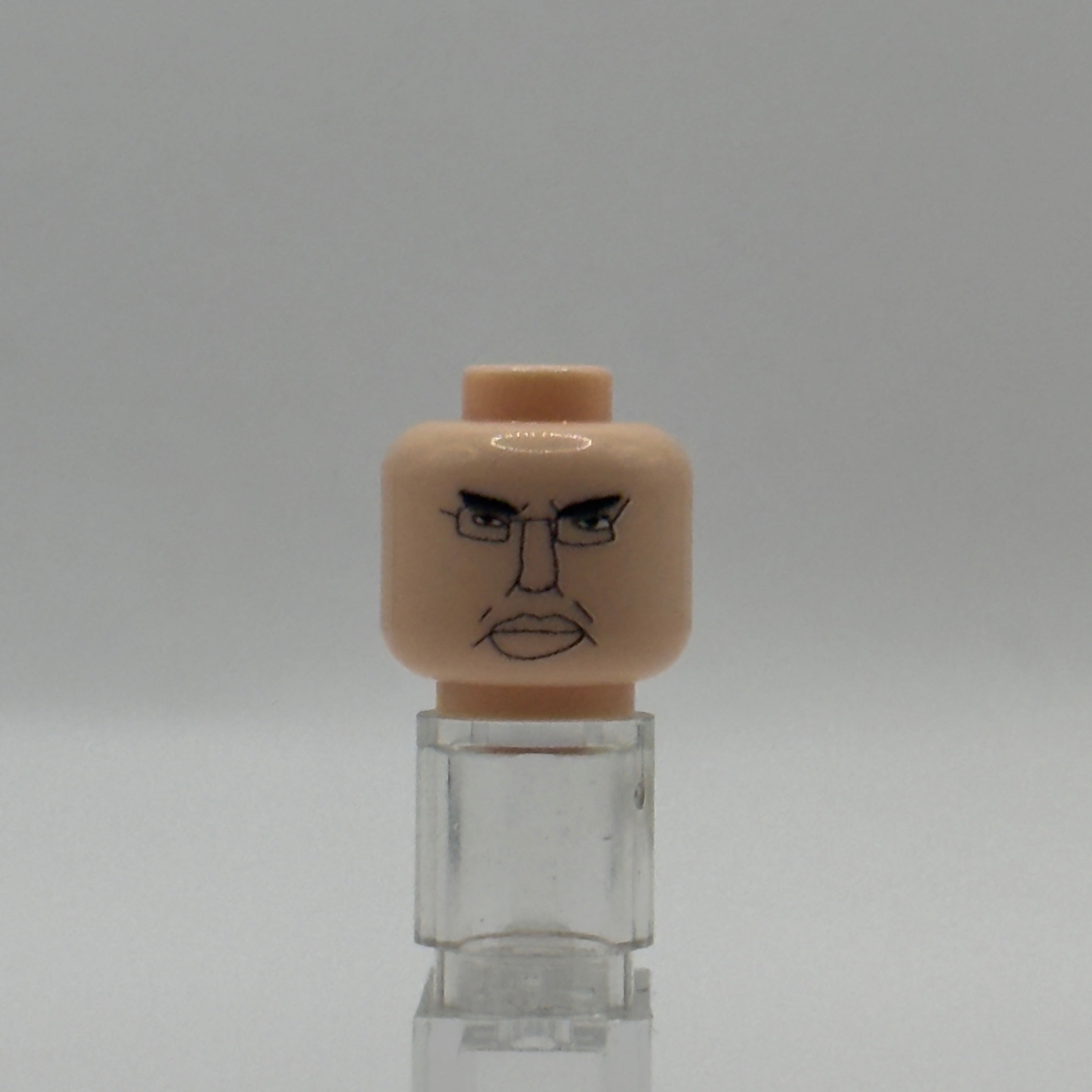 A beige plastic toy head with a printed angry face, displayed on a clear acrylic stand against a neutral gray background.